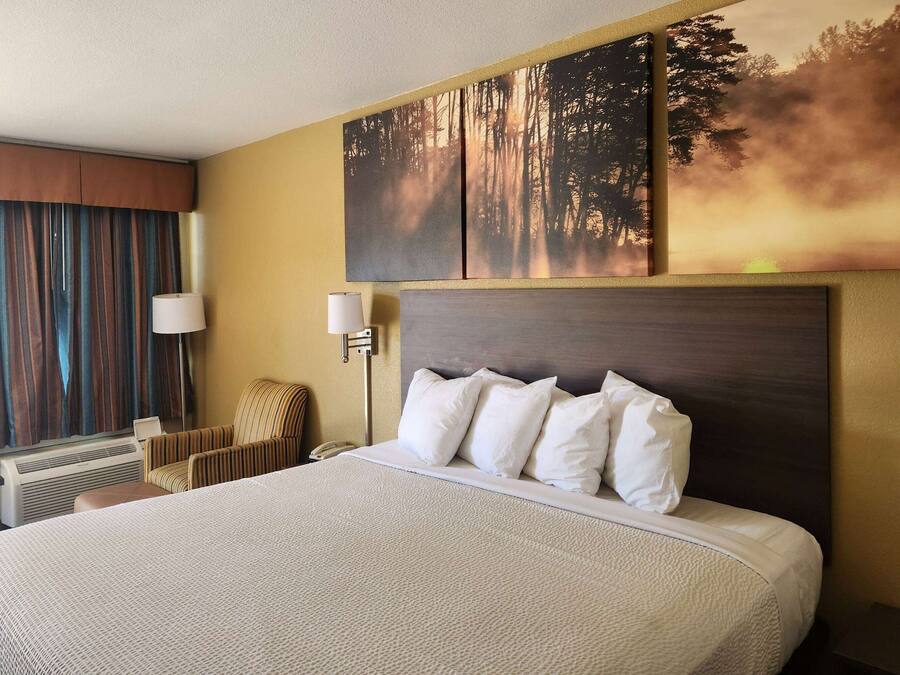 A hotel room with a large bed, a headboard, a wall-mounted artwork, a window with curtains, a chair, and a desk with a lamp.