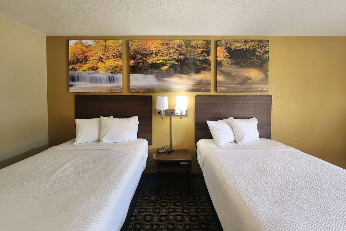 Habitación de hotel con dos camas, una mesita de noche con lámparas y un cuadro colgado en la pared de una cascada.