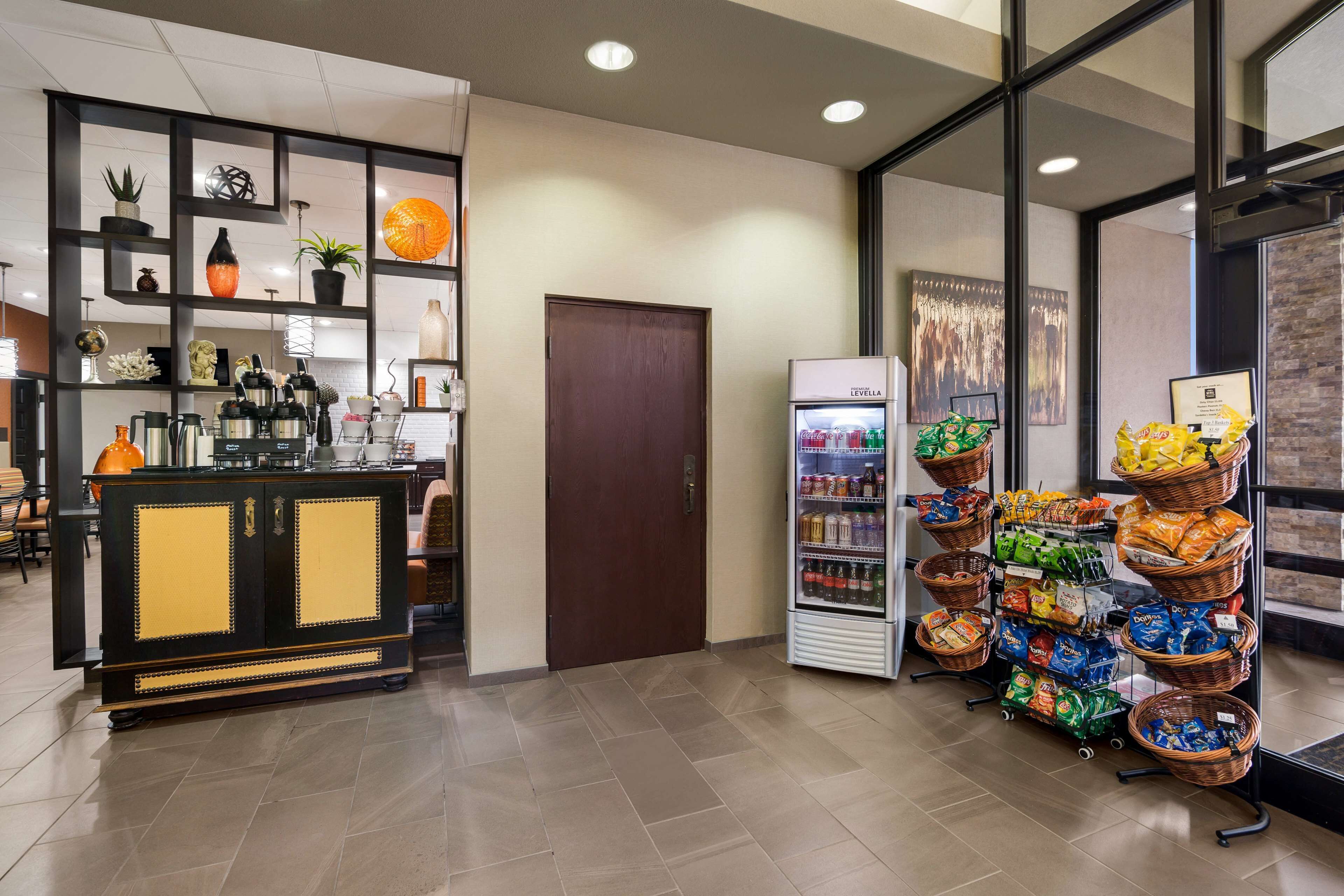 Un vestíbulo de hotel con una recepción, un refrigerador lleno de bebidas y una exhibición de aperitivos y frutas.