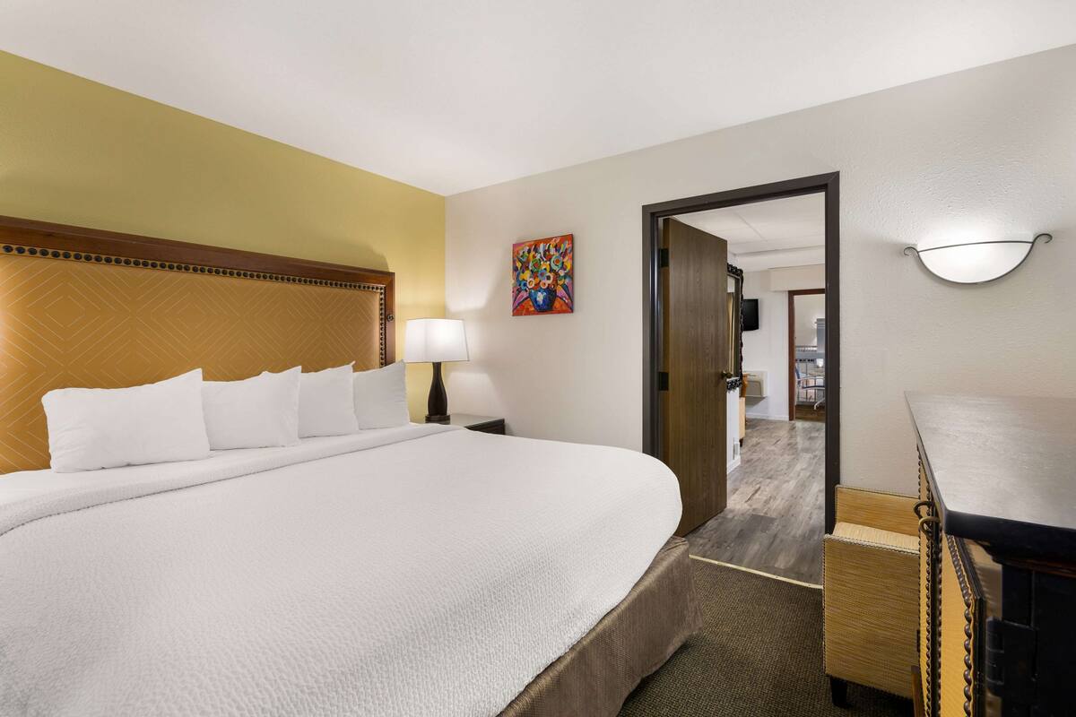 A hotel room with a large bed, a wooden headboard, a bedside table with a lamp, a colorful artwork on the wall, and a door leading to another room.