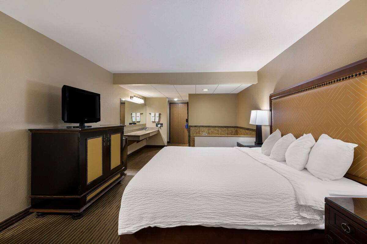 A hotel room with a bed, a TV, a desk, and a bathroom visible through an open door.