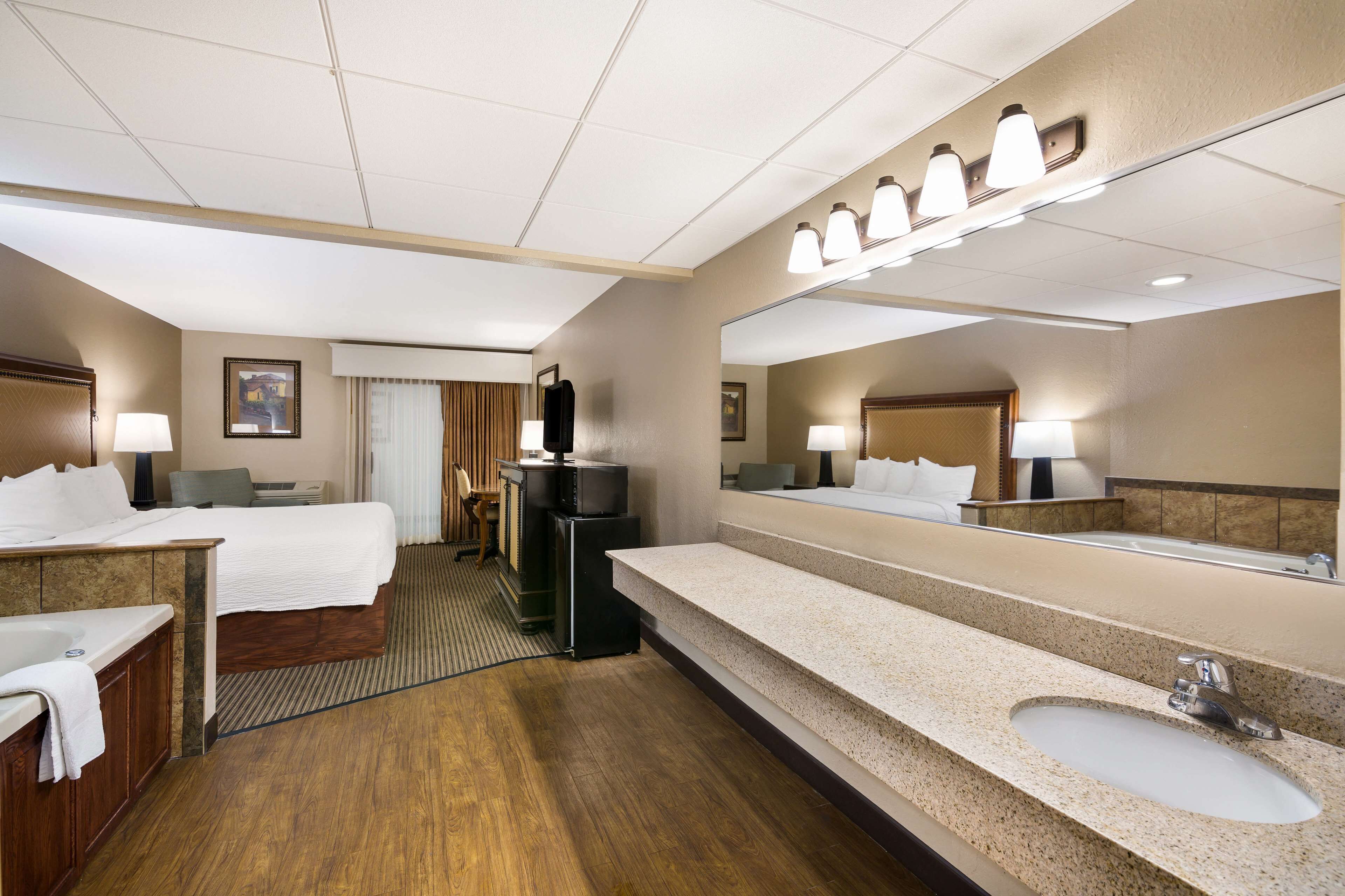 Une chambre d’hôtel avec un grand lit, un coin salon comprenant un fauteuil et une petite table, une télévision, un grand miroir et une salle de bain moderne équipée d’un lavabo et de toilettes.