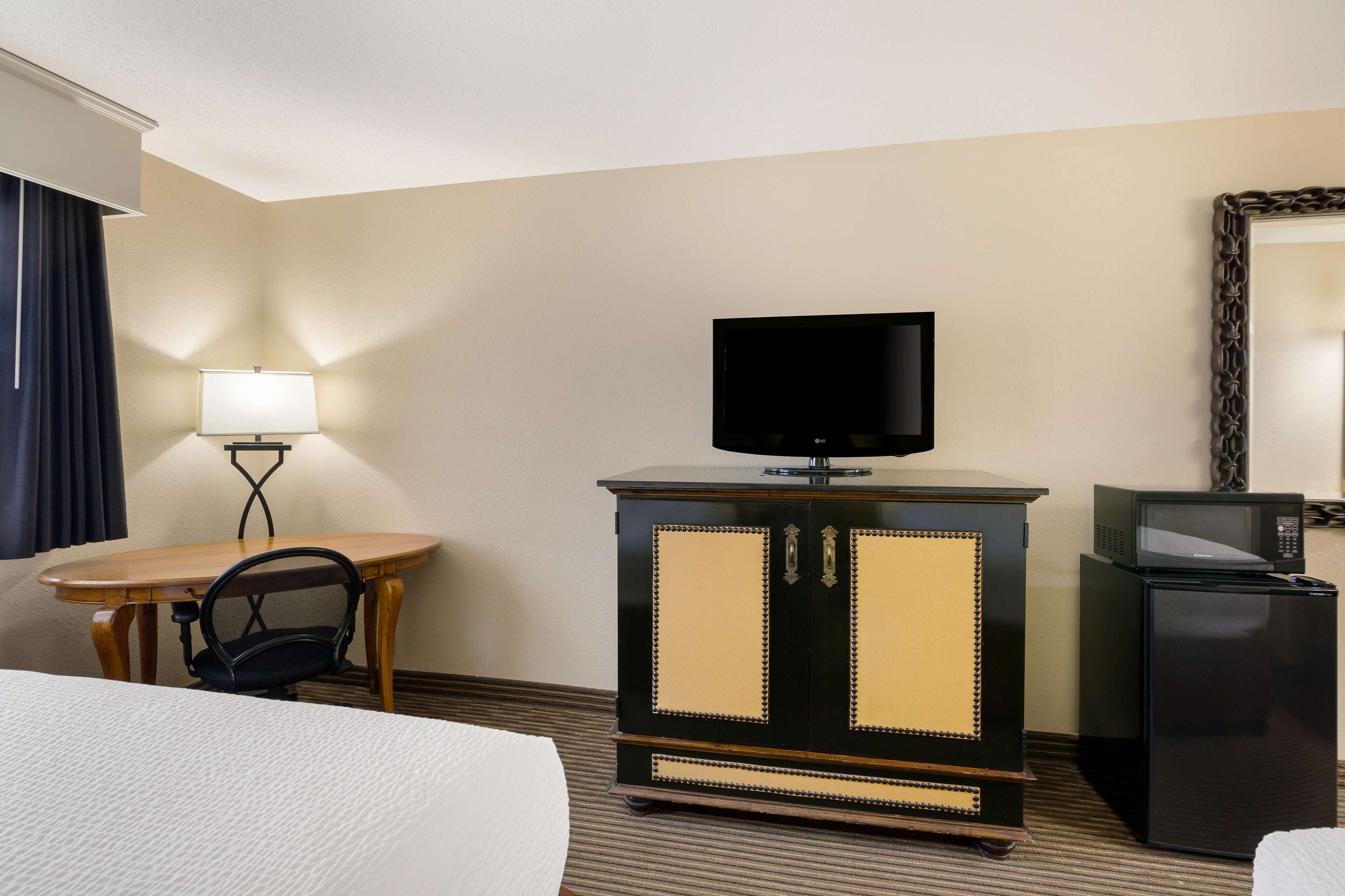 Habitación de hotel con cama, un escritorio de madera con silla, televisor en un mueble, microondas y un espejo en la pared.