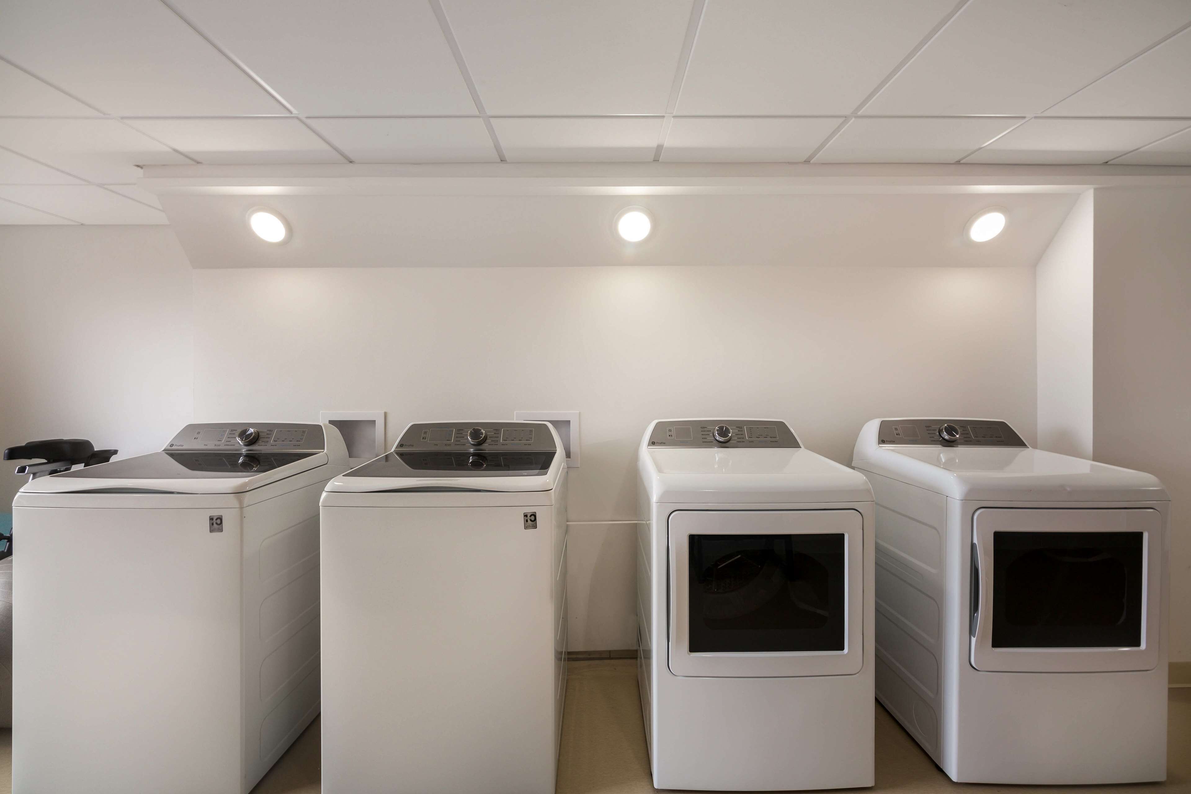 A laundry room with four white front-loading washers and dryers, a white ceiling with recessed lighting, and a plain white wall.