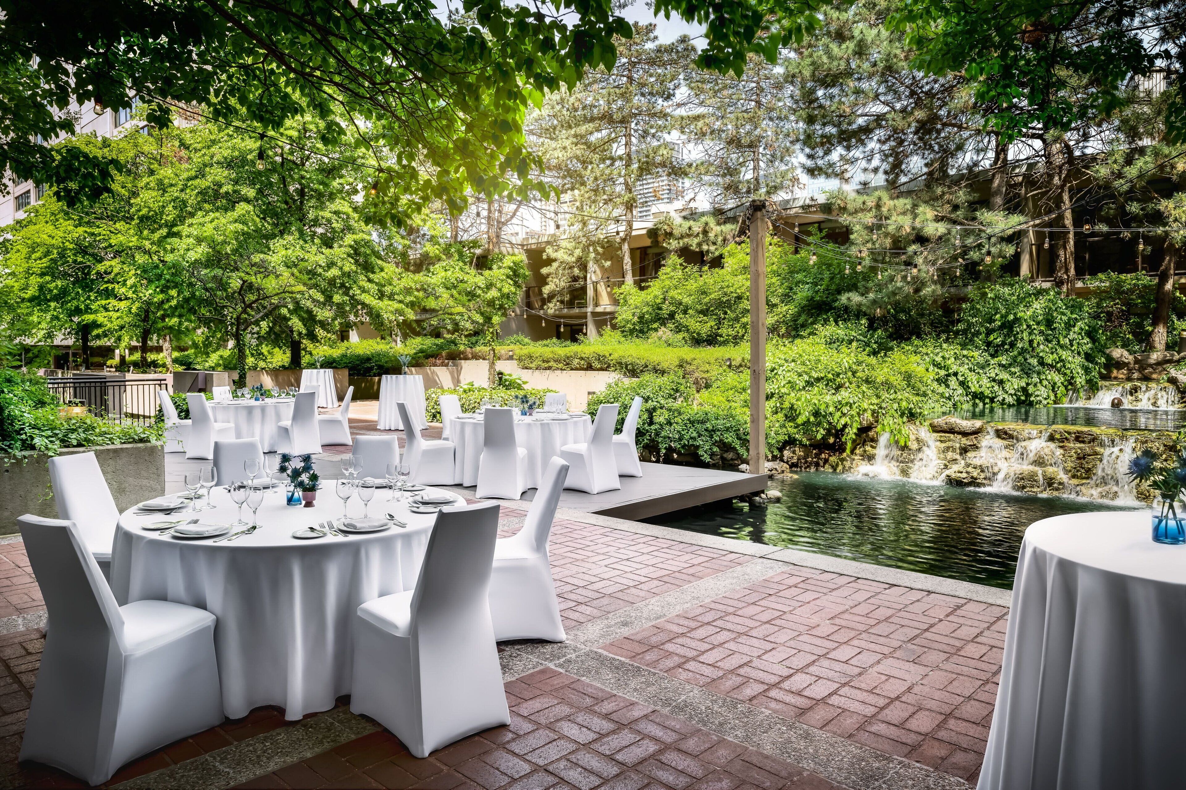 An outdoor dining area with round tables and white chairs, surrounded by lush greenery and a small pond.