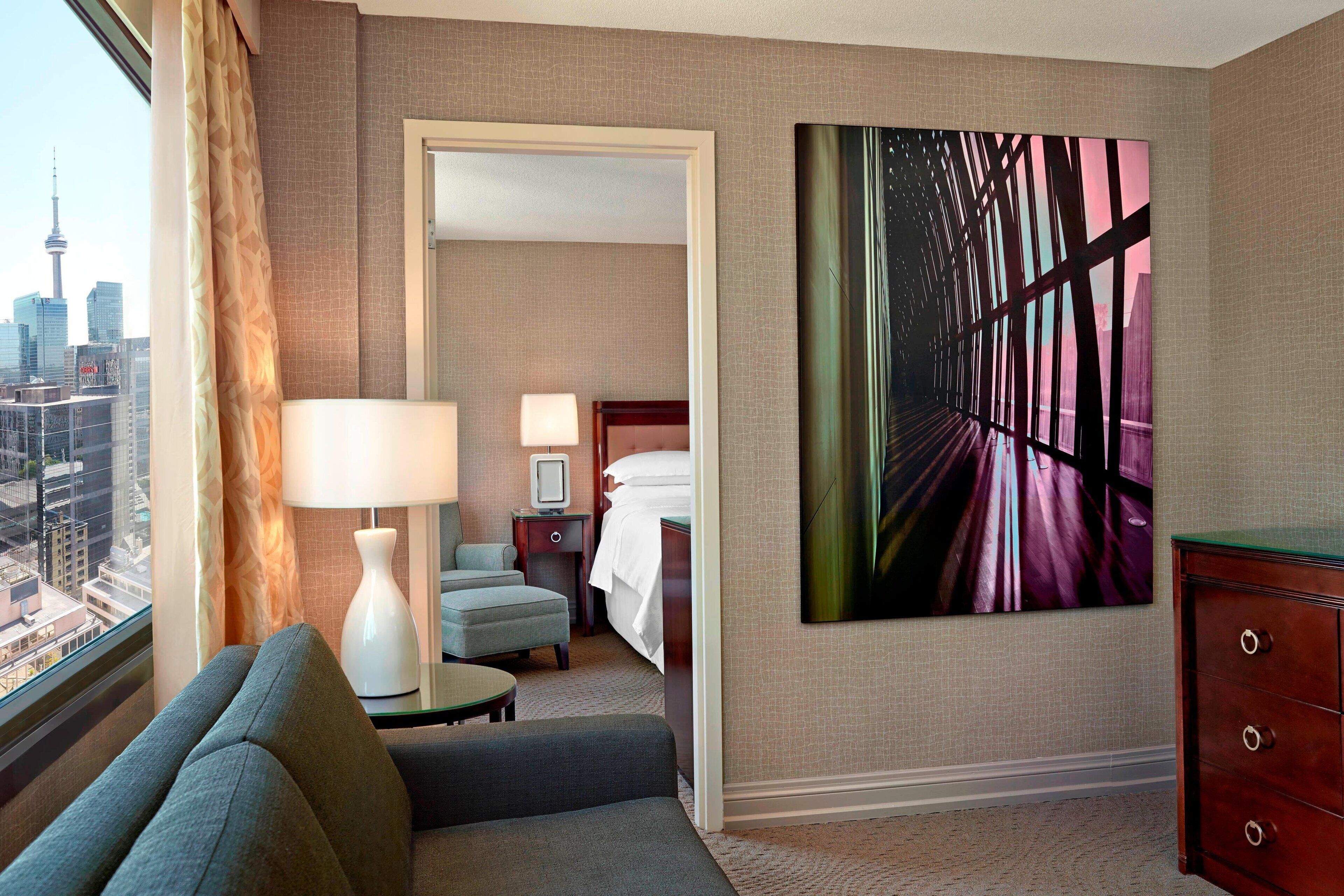 Een hotelkamer met uitzicht op de stadshemel, een bed, een bureau met lamp, een stoel en een groot abstract schilderij aan de muur.