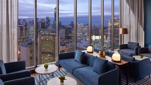 Habitación de hotel moderna con amplia vista de la ciudad, un sofá azul, dos mesas redondas y una lámpara.