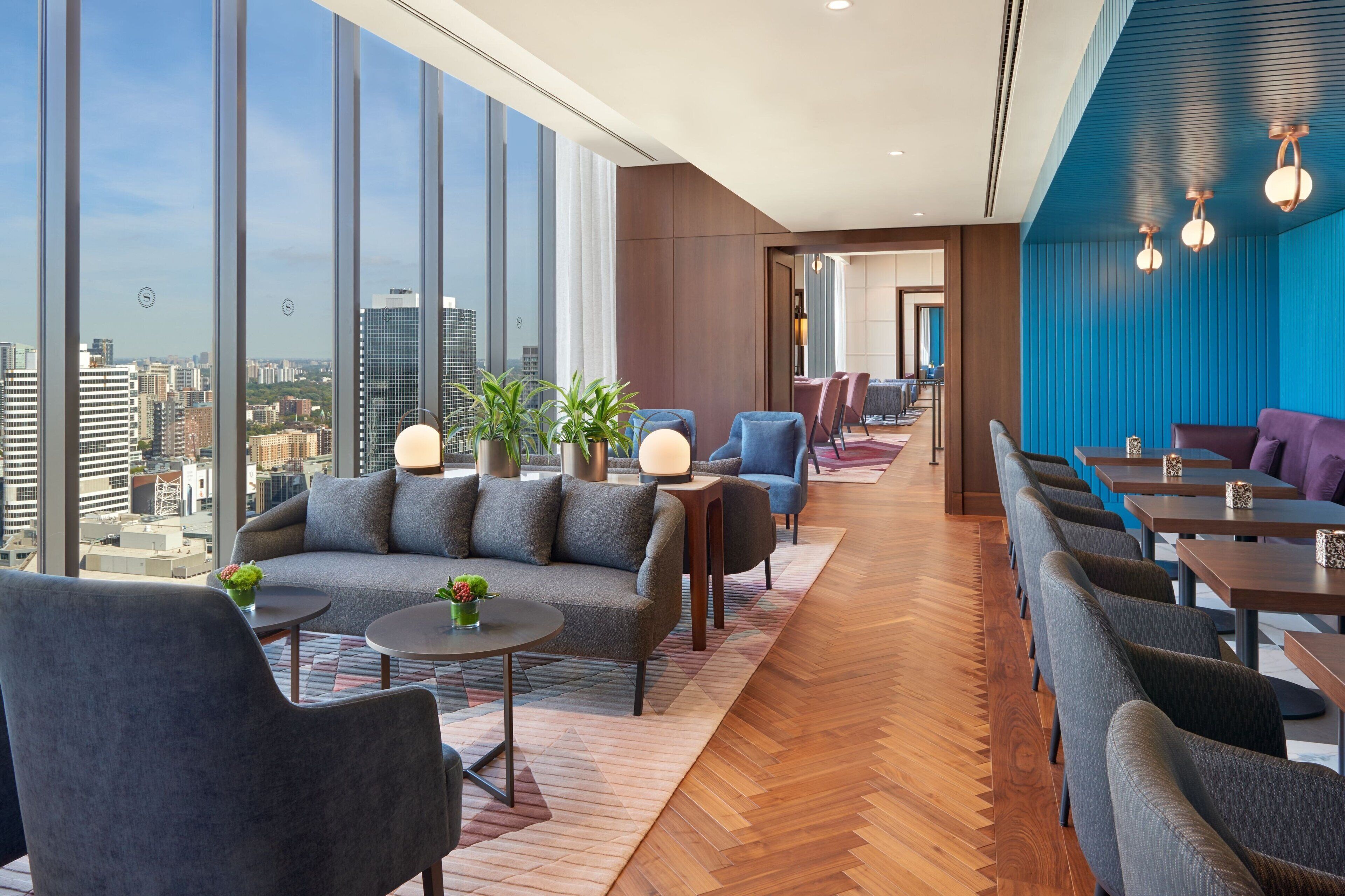 A modern hotel lobby with a high ceiling, large windows offering city views, and a mix of seating areas including sofas and tables.