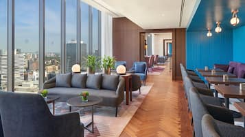A modern hotel lobby with a high ceiling, large windows offering city views, and a mix of seating areas including sofas and tables.