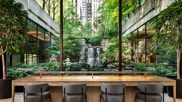 Un espacio interior moderno con una mesa de madera larga, tres sillas y vistas a una cascada y follaje a través de amplios ventanales.