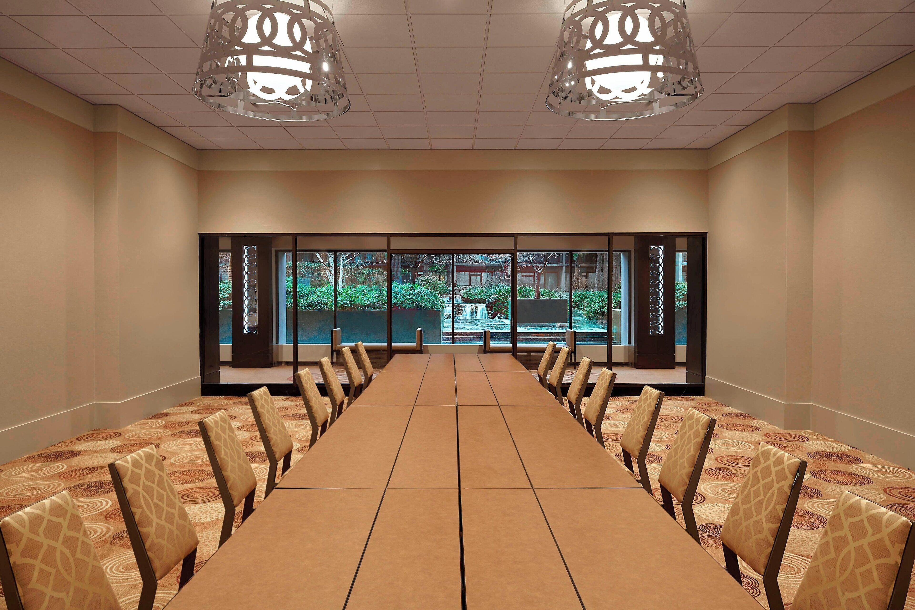 Un salón de conferencias con una larga mesa y sillas, ventanas amplias con vistas a zonas verdes y luminarias decorativas en el techo.