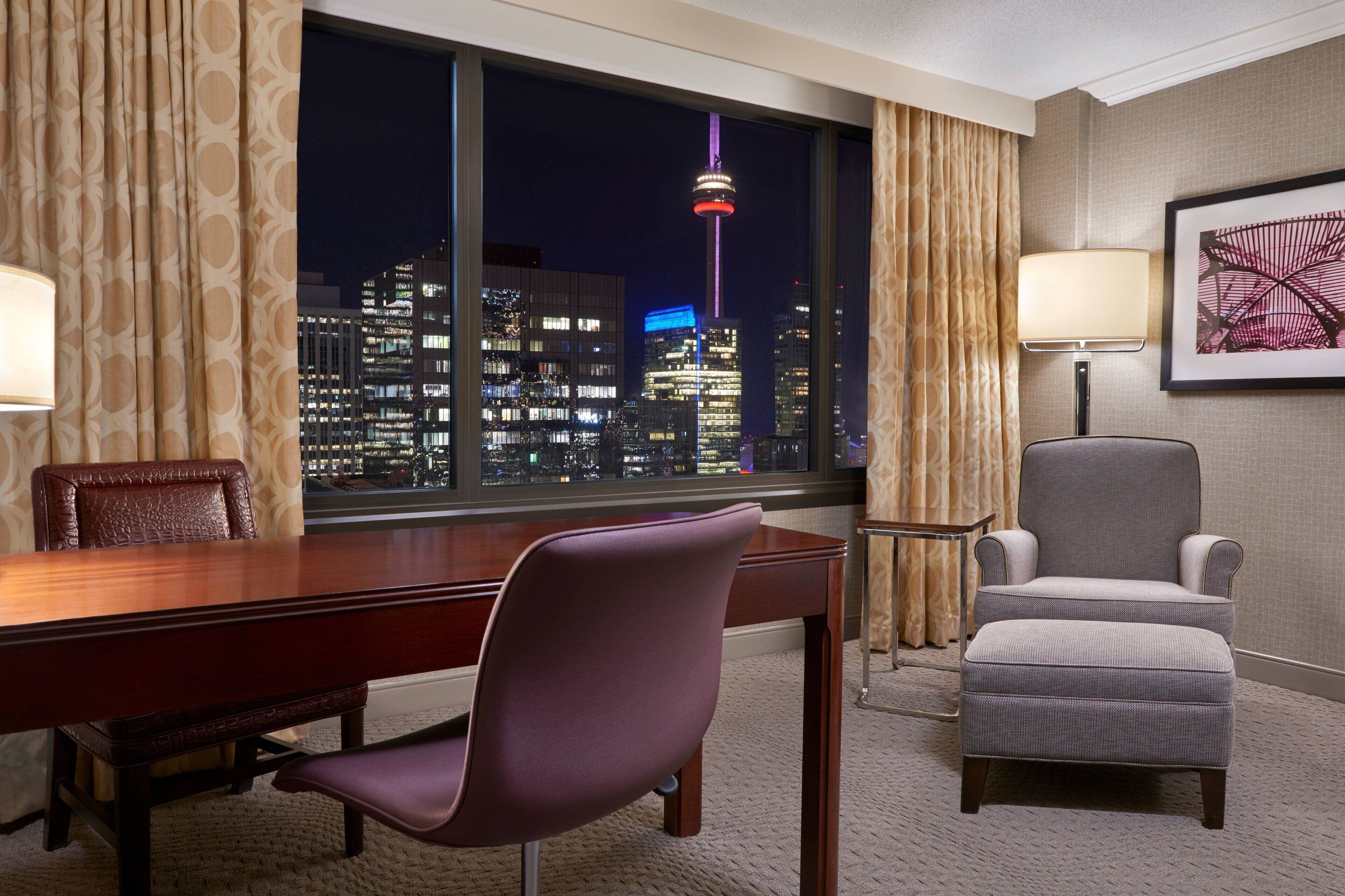 A hotel room with a large window offering a nighttime city view, a wooden desk, a purple chair, a grey armchair, and a lamp.