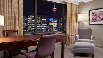 A hotel room with a large window offering a nighttime city view, a wooden desk, a purple chair, a grey armchair, and a lamp.