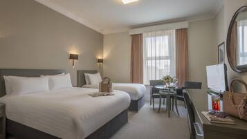 A hotel room with a large bed, a smaller bed, a desk with a chair, a round table, a mirror, and a window with curtains.
