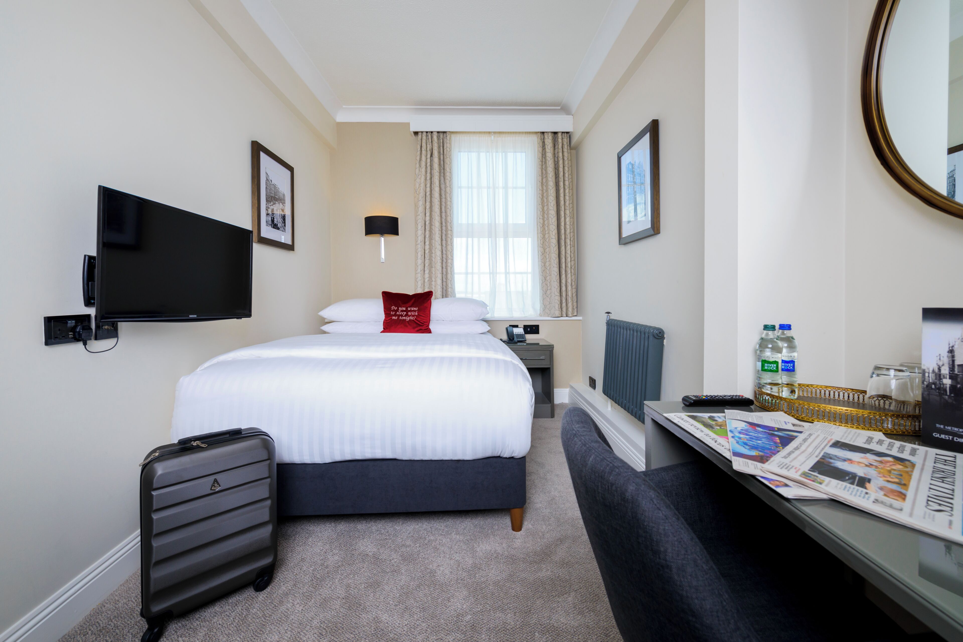 A hotel room with a bed, a TV, a desk with a newspaper, a suitcase, and a window with curtains.