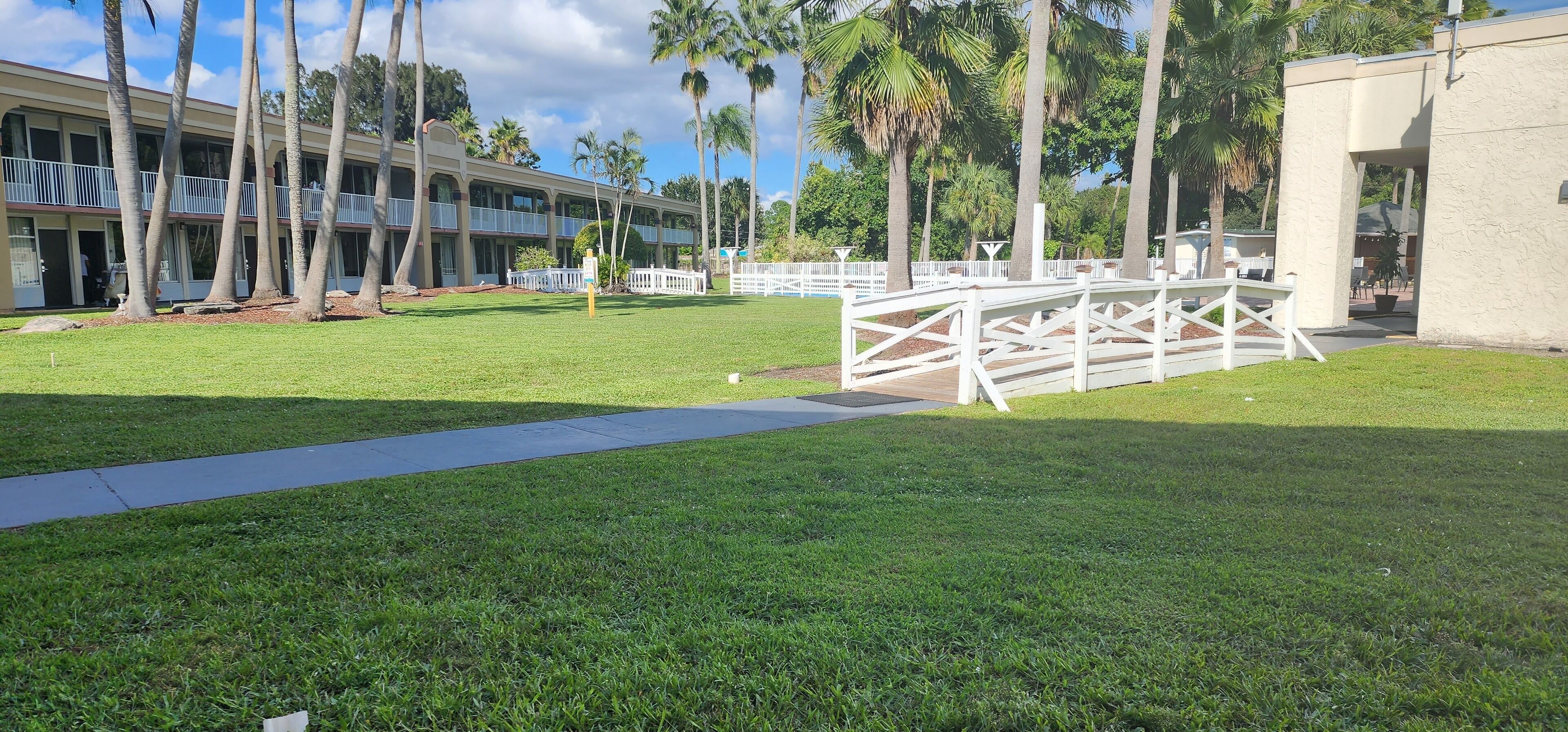 Un patio de hotel con un camino pavimentado, palmeras y una valla blanca.