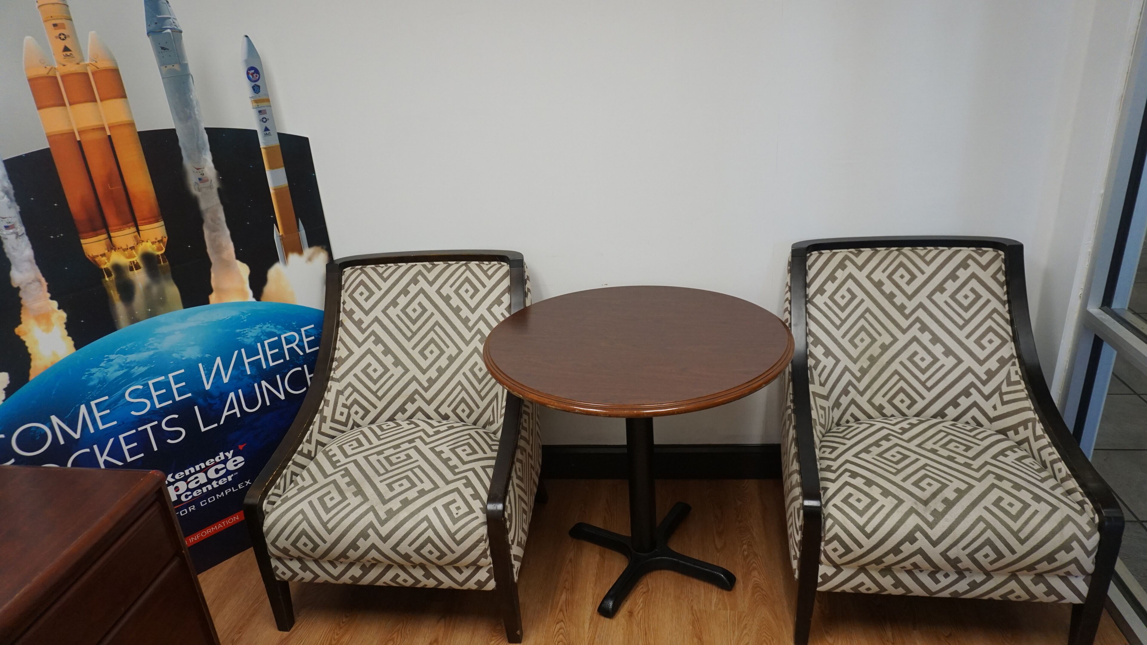 A small room with two patterned chairs and a round table. A poster with rocket images and text is visible on the wall.