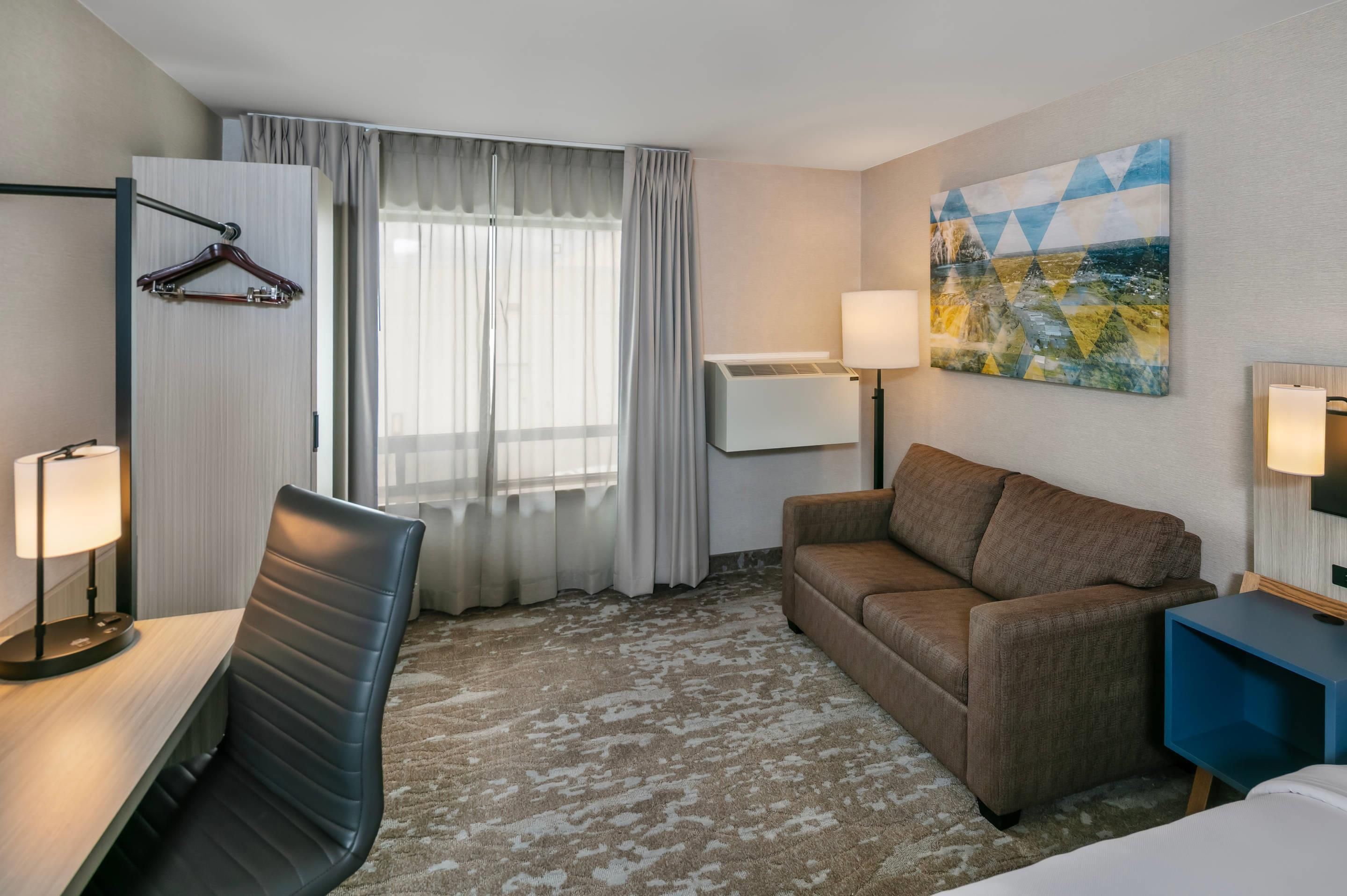 A hotel room with a bed, a desk, a chair, a sofa, a lamp, a TV, and a painting on the wall.