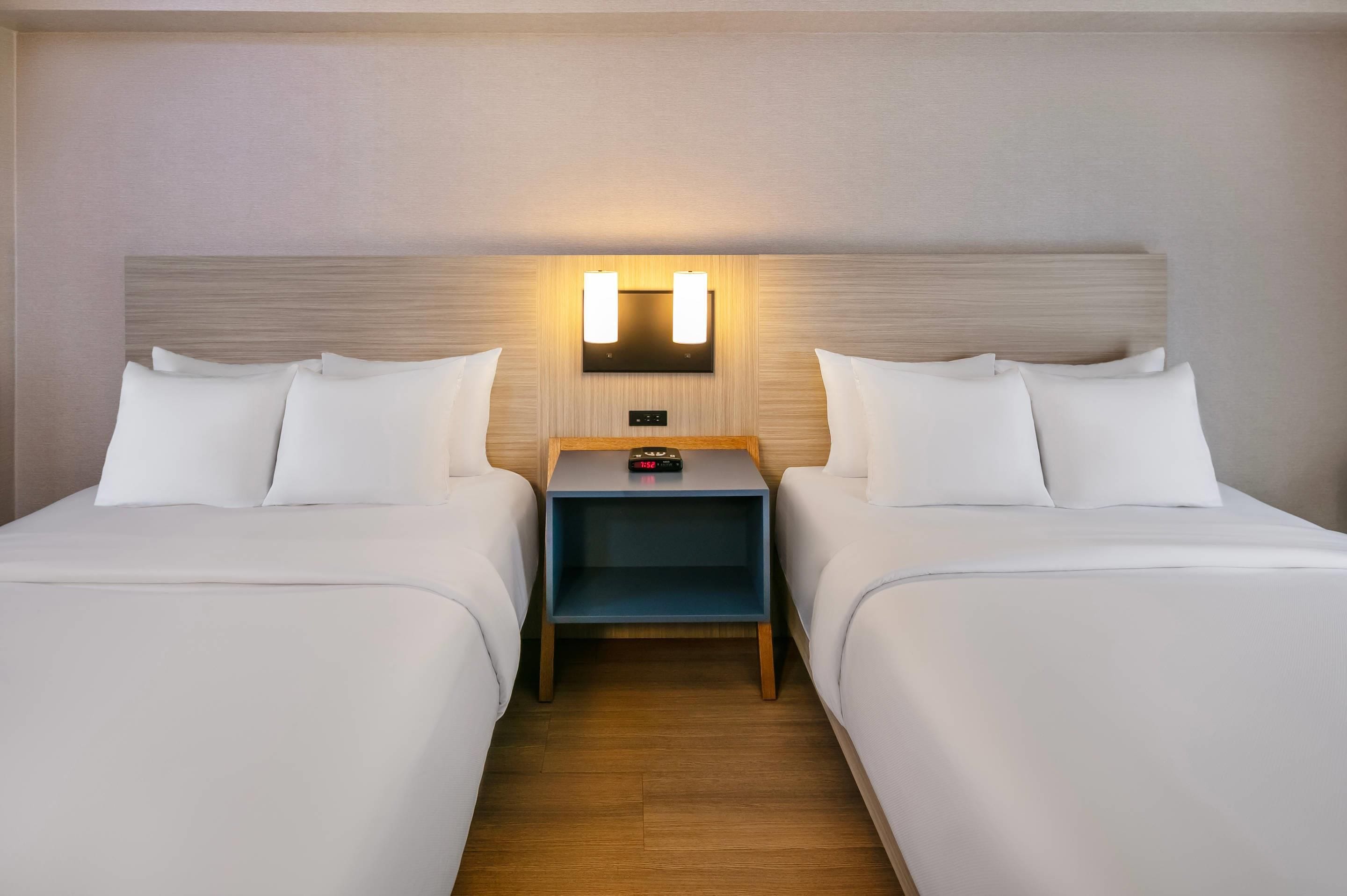 A hotel room with two beds, a nightstand, a wall-mounted lamp, and a digital clock.