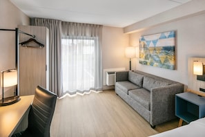 A hotel room with a grey sofa, a desk with a lamp, a chair, a painting on the wall, and a window with curtains.