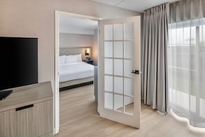 A hotel room with a bed, a TV, a nightstand, and a door leading to another room.
