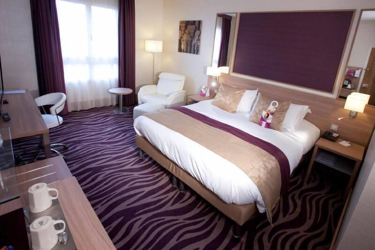 A hotel room with a bed, a desk, a chair, a small table, and a nightstand with a lamp.