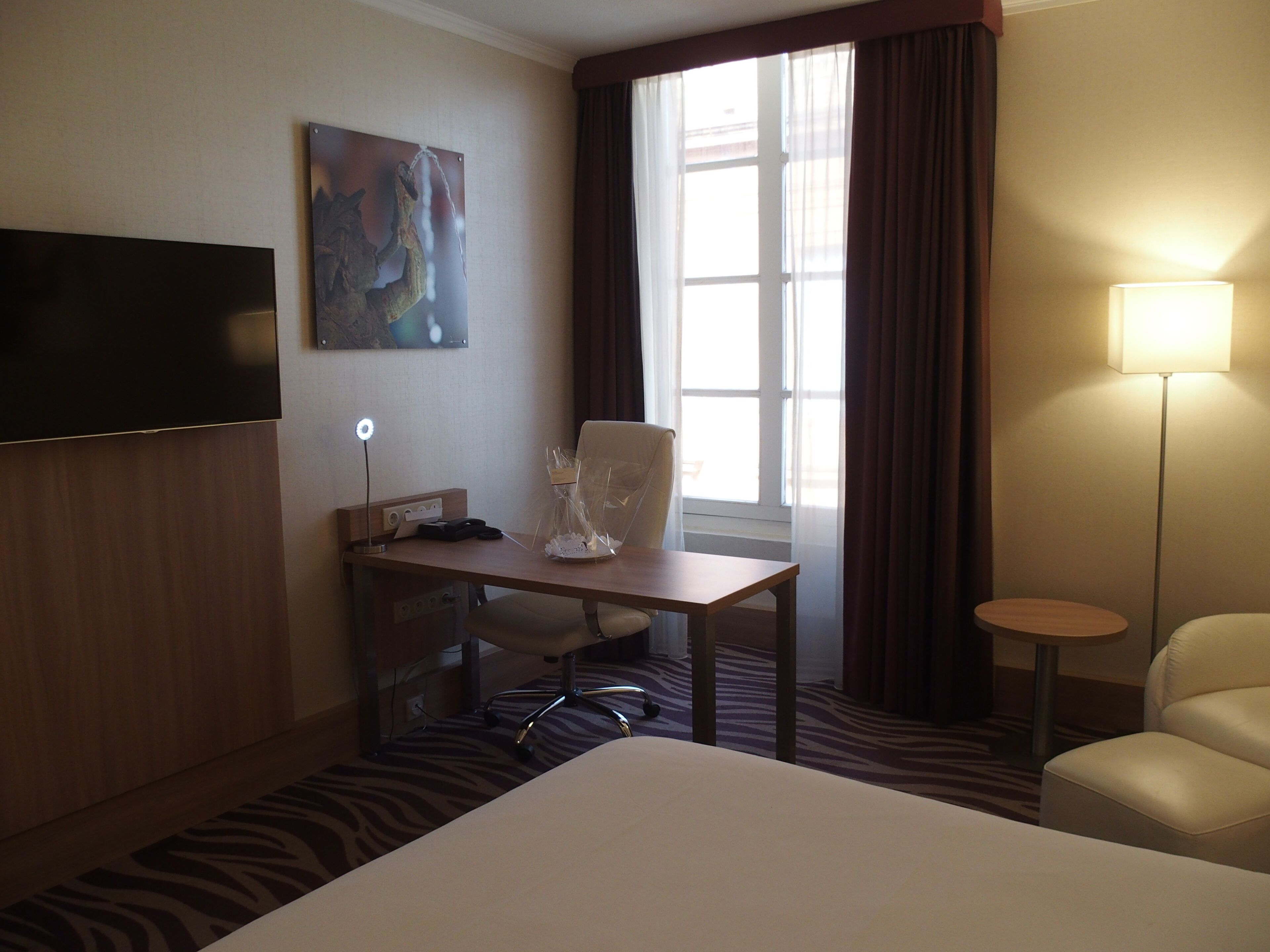 A hotel room with a bed, a desk with a chair, a television, a lamp, and a window with curtains.