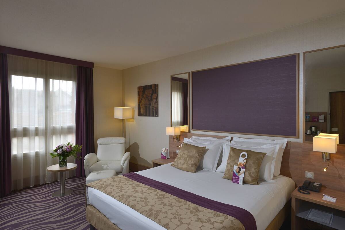 A hotel room with a large bed, a chair, a small table, and a nightstand.
