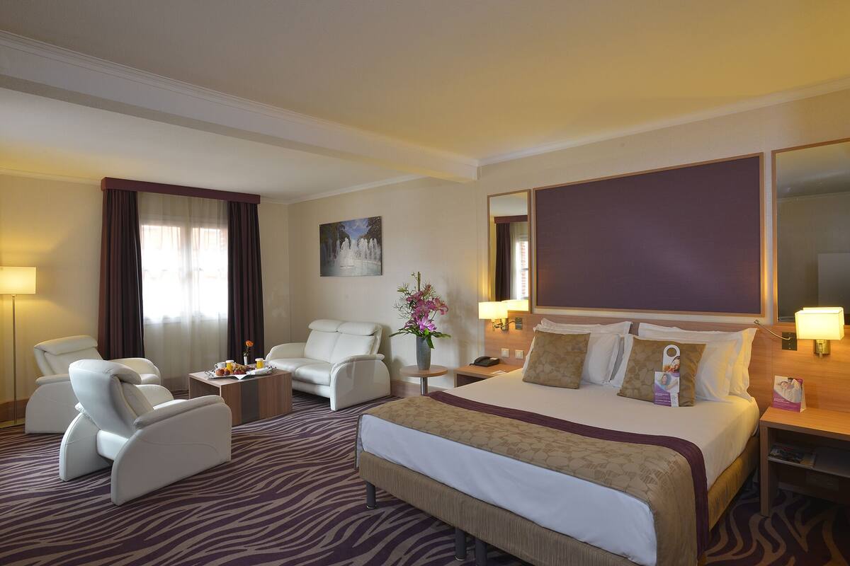 A hotel room with a large bed, two white armchairs, a small table, a vase of flowers, and a painting on the wall.
