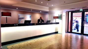 A hotel reception area with a receptionist behind the counter, a large abstract painting on the wall, and a glass door leading outside.