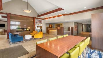 A spacious hotel lobby with a long wooden reception desk, yellow and blue seating, and a large dining area.
