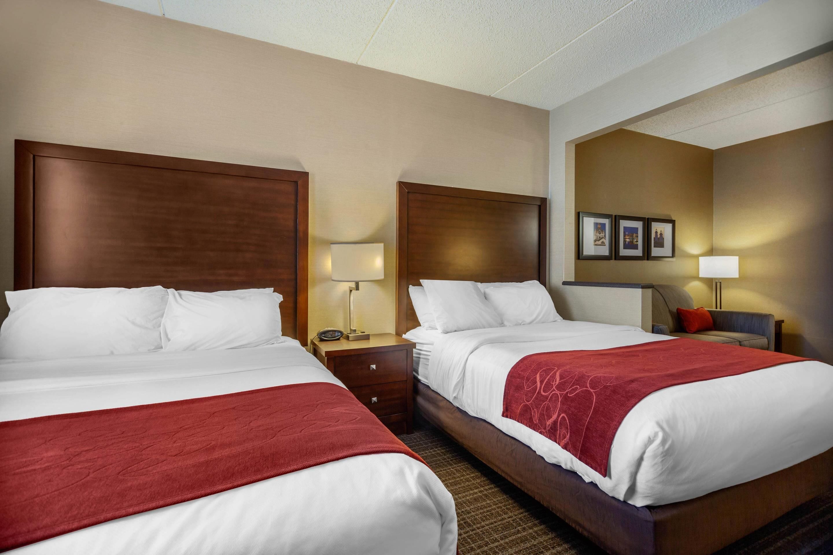 A hotel room with two beds, each with a wooden headboard, white bedding, and a red throw blanket. There are bedside tables with lamps, a wall-mounted TV, and framed pictures.