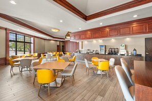 Un grand espace à manger doté d’un plancher en bois, de chaises jaunes et grises, et d’un coin cuisine équipé d’une machine à café.