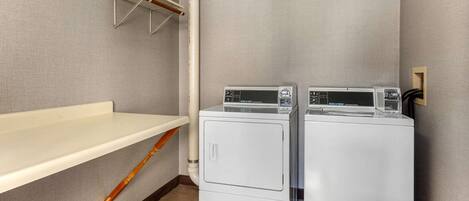 A laundry room with two stacked washers and dryers, a wooden drying rack, and a white shelf.