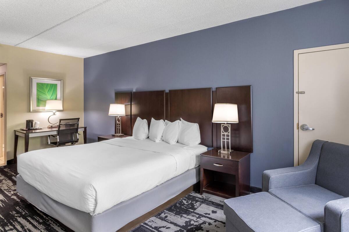 A hotel room with a large bed, two bedside tables with lamps, a desk with a chair, and a grey armchair.
