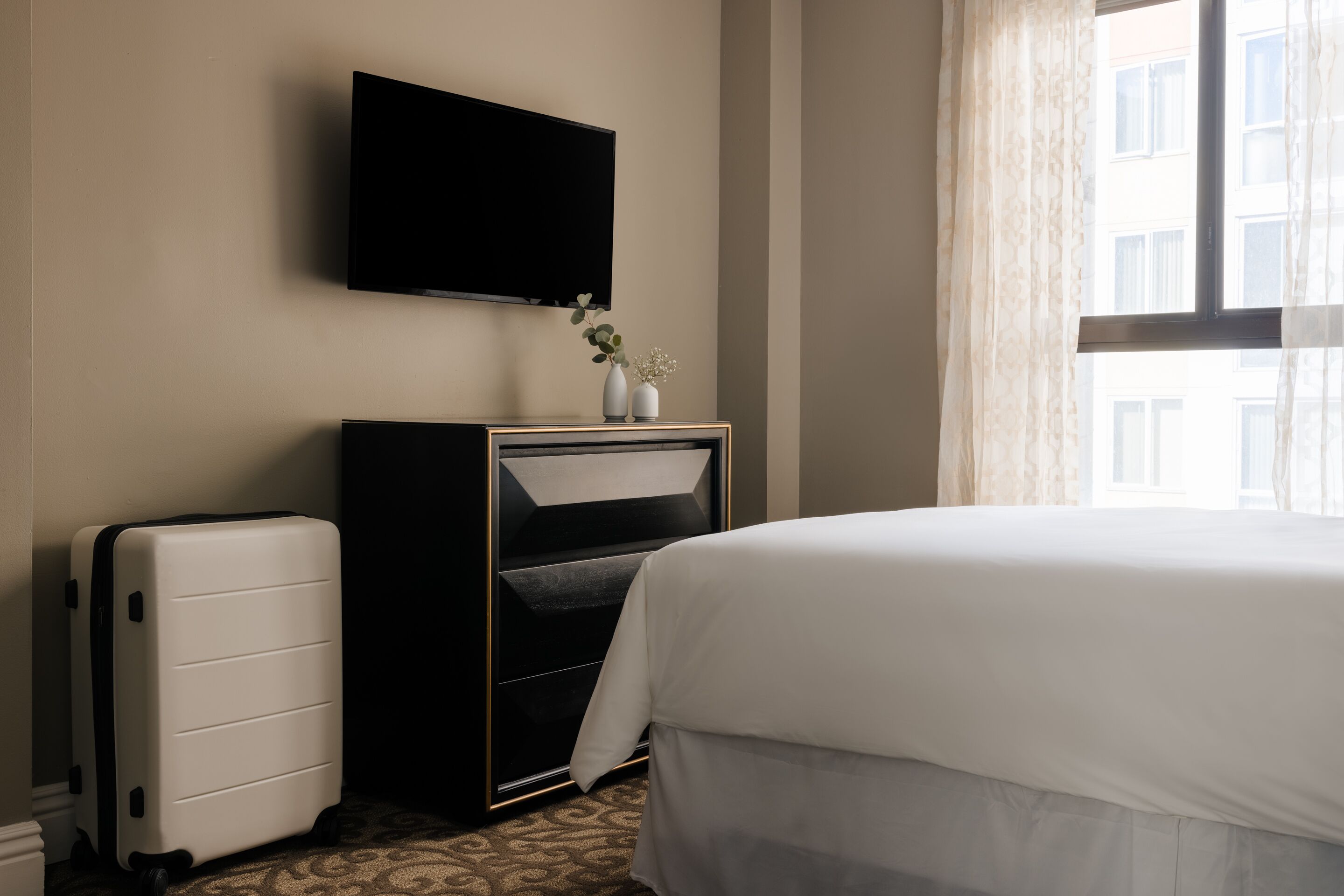 A hotel room with a bed, a flat-screen TV mounted on the wall, a desk with a computer, and a suitcase.
