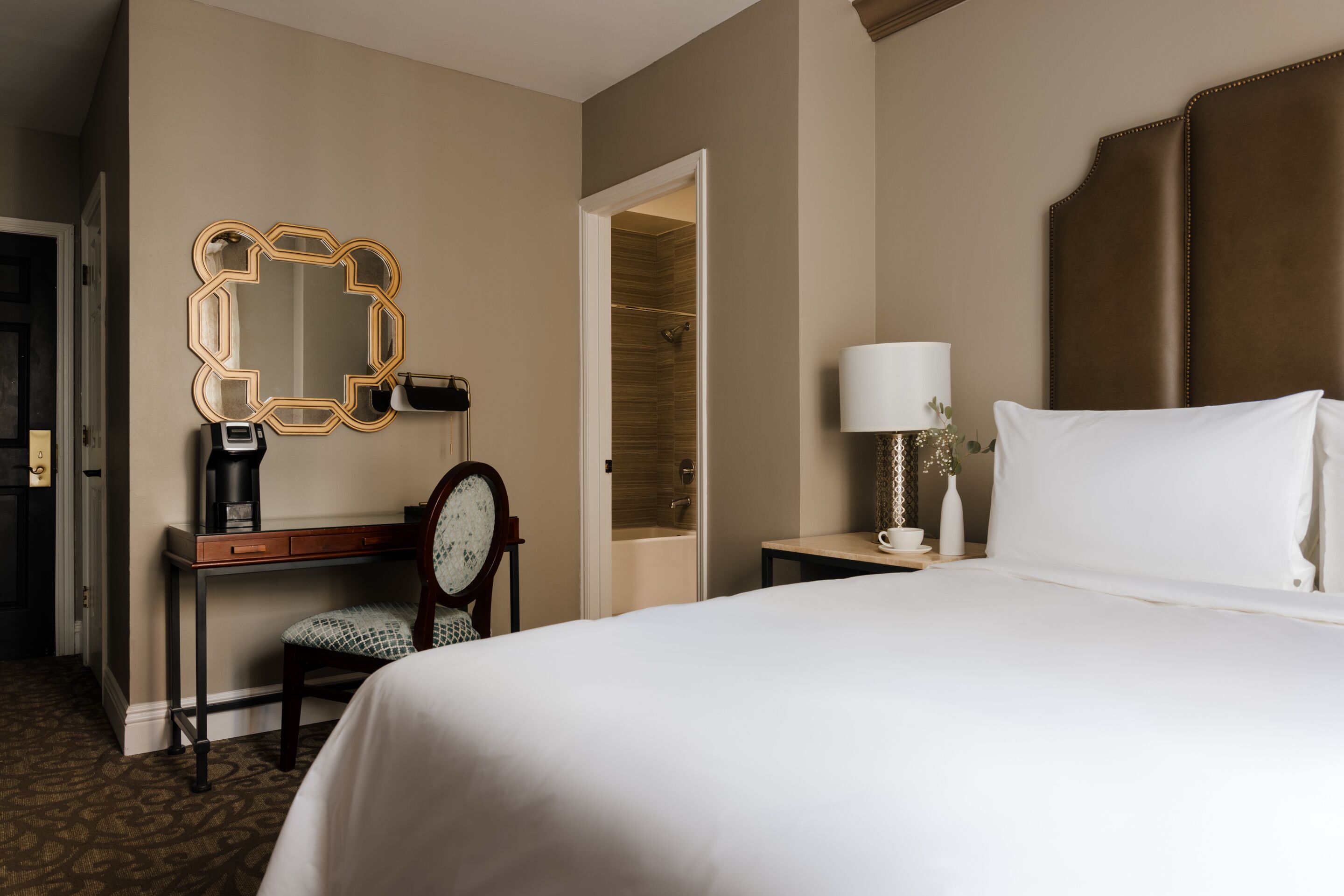 Una habitación de hotel con una cama grande, un escritorio con silla, un espejo, una mesita de noche con lámpara y un vestidor.