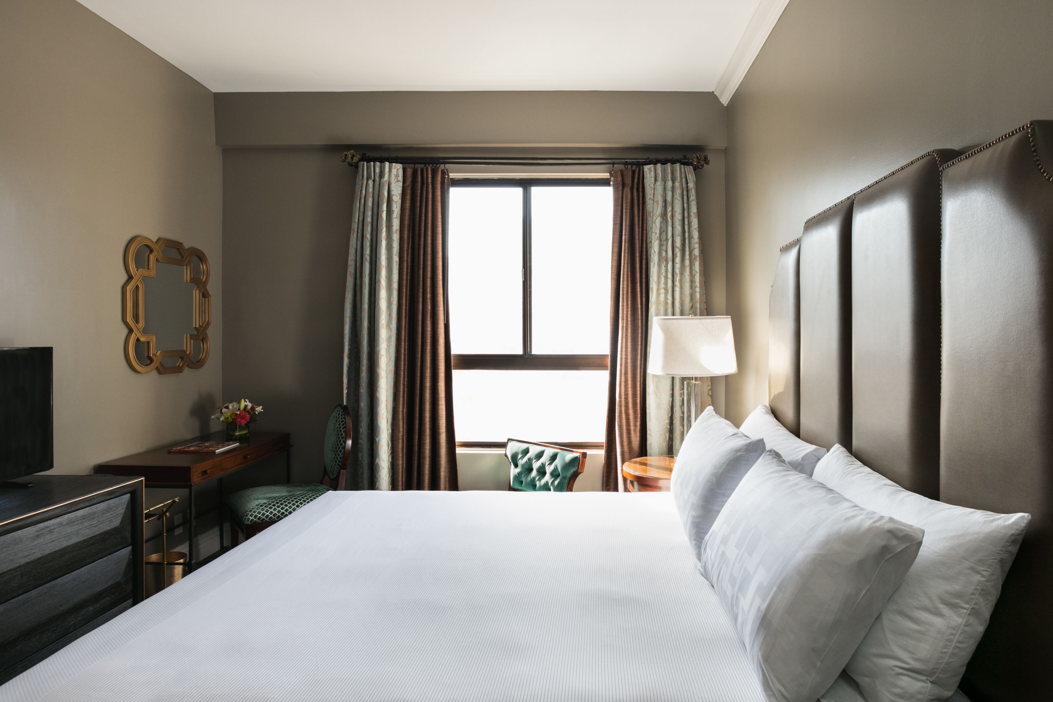 A hotel room with a large bed, a desk, a chair, a TV, and a window with curtains.