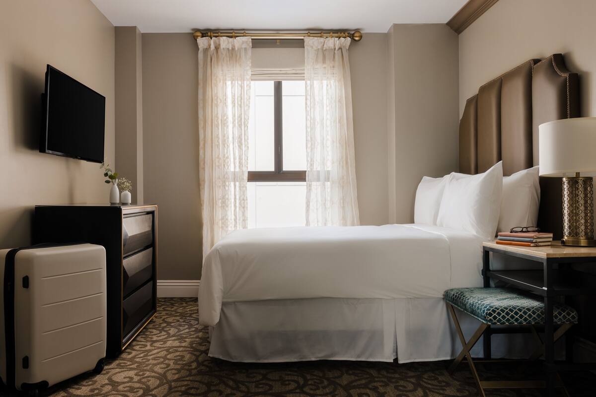 A hotel room with a bed, a desk with a lamp, a television, a dresser, and a suitcase.