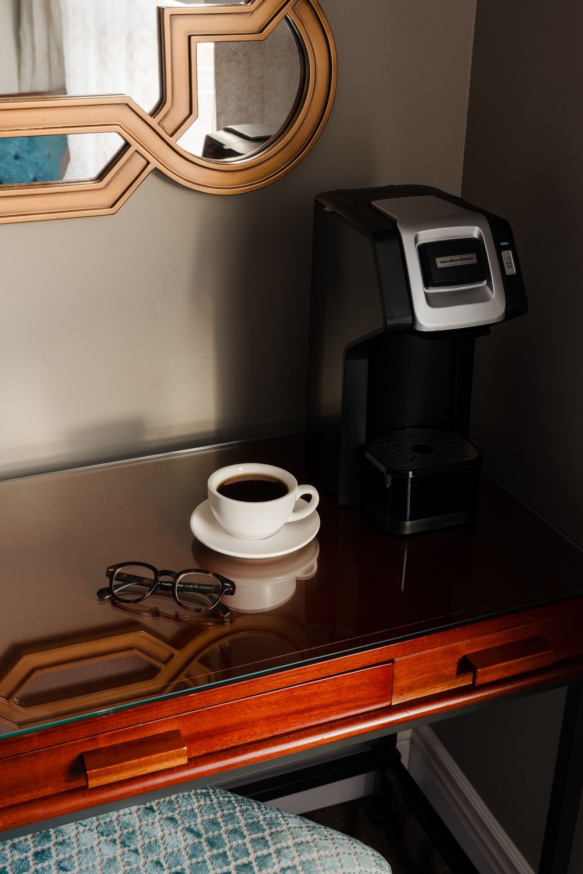 A coffee maker on a wooden table with a cup of coffee and a pair of glasses.