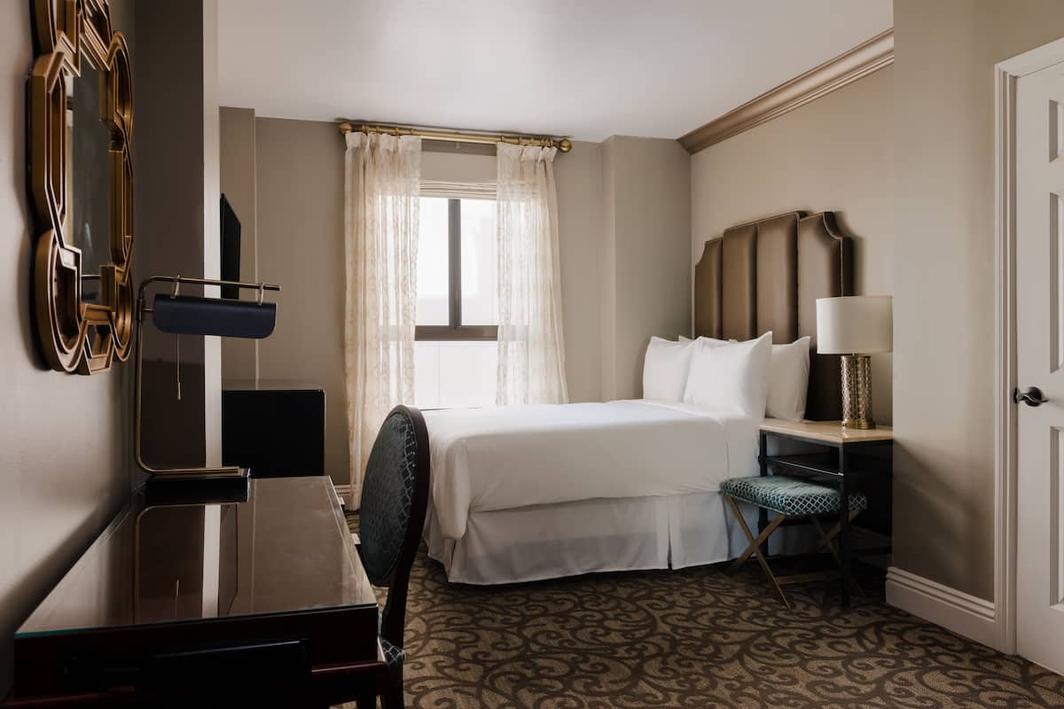 A hotel room with a bed, a desk, a chair, a lamp, and a mirror.