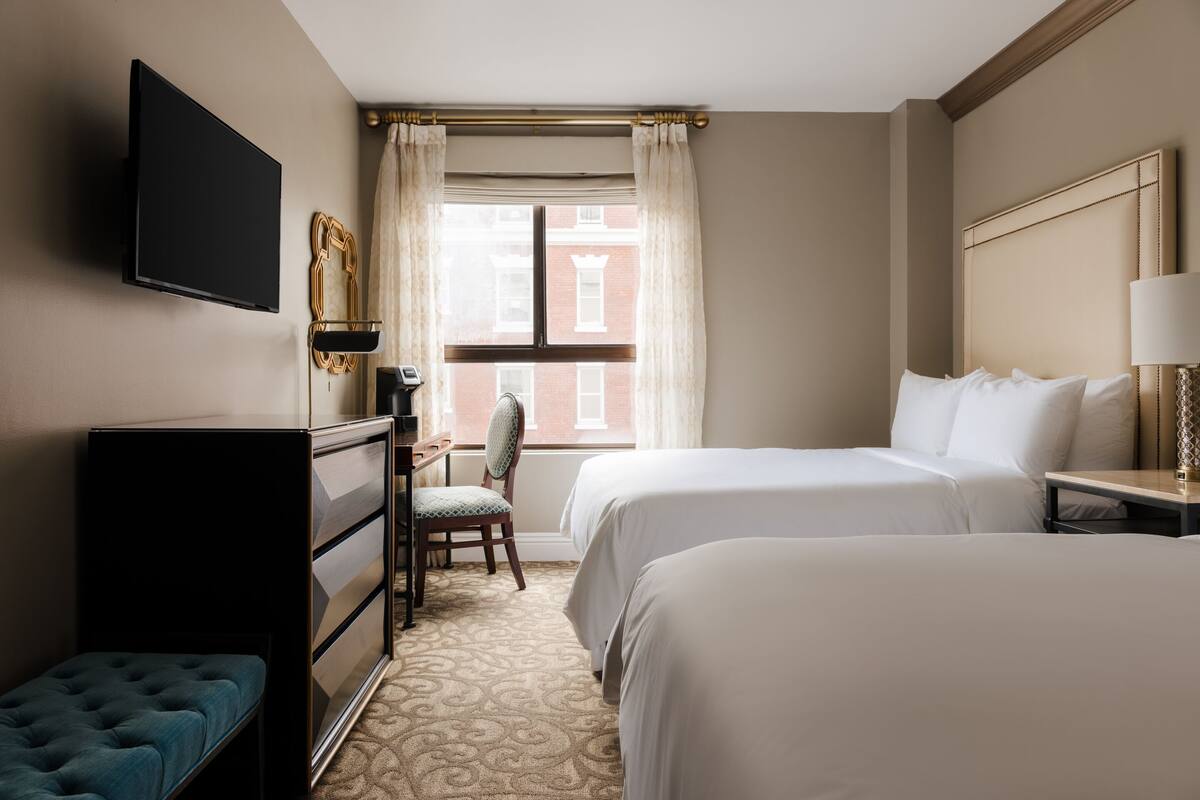 A hotel room with two beds, a desk, a chair, a television, and a window with curtains.