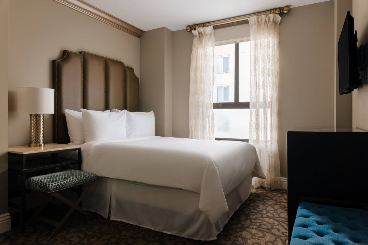 A hotel room with a large bed, a desk with a lamp, a chair, and a TV mounted on the wall.