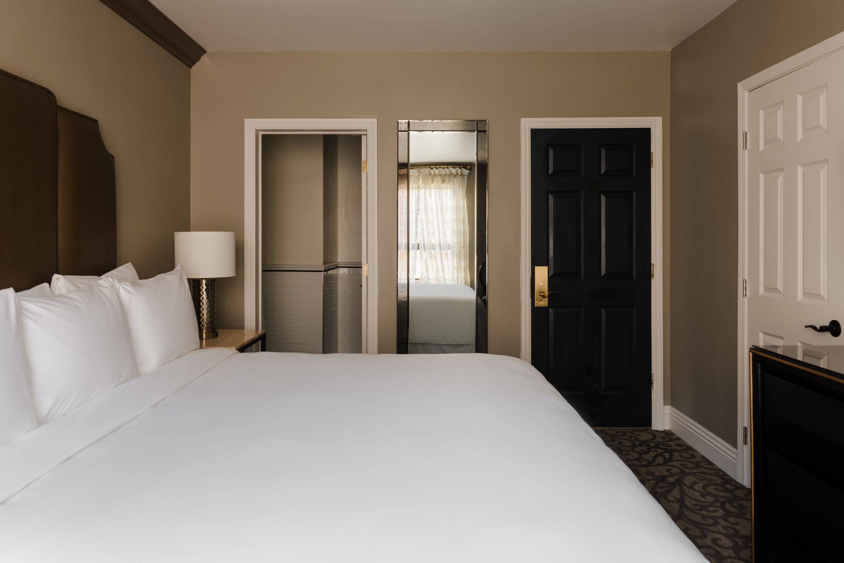 A hotel room with a large bed, a bedside table with a lamp, a door leading to a bathroom, and a door to another room.