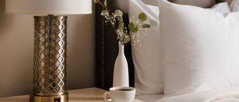 A neatly made bed with white linens, a bedside table with a lamp, and a vase with flowers.
