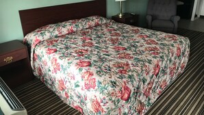 Una habitación de hotel con cubrecama floral, una mesita de noche con lámpara, una silla y alfombra a rayas.
