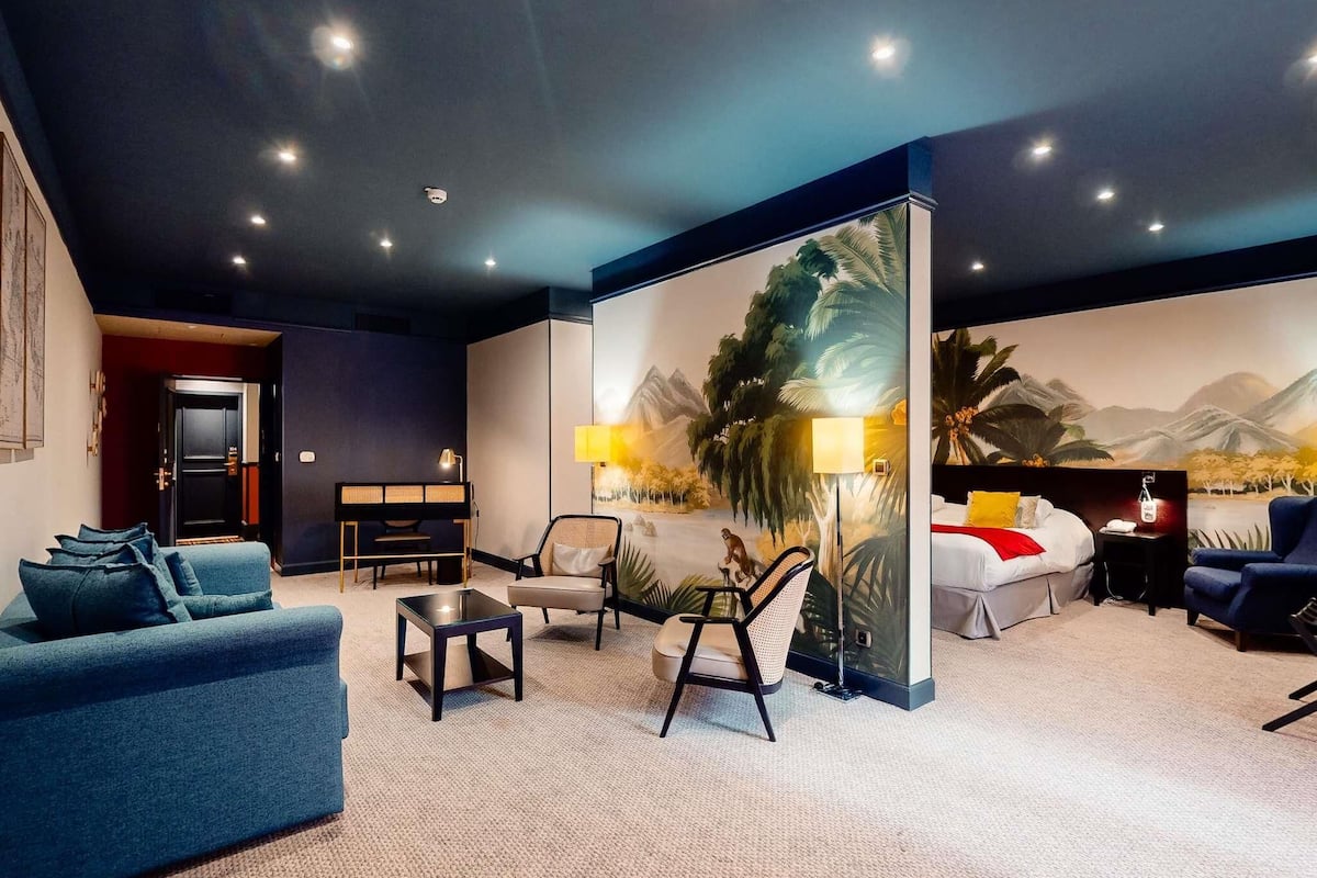 A modern hotel room with a sofa, armchair, and a bed with a mountain view mural.