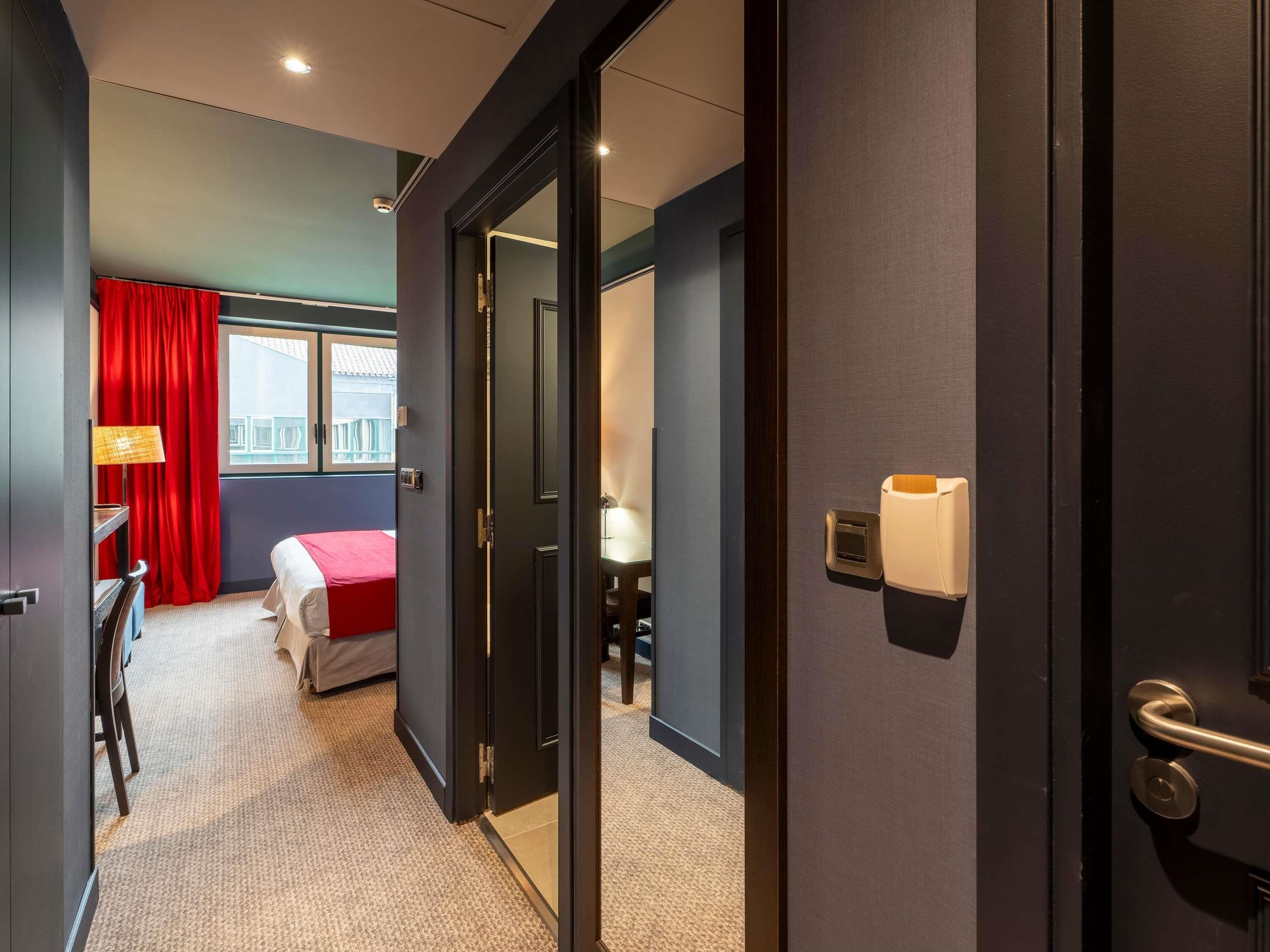 Une chambre d’hôtel avec un lit, un bureau, une lampe et une fenêtre avec des rideaux rouges.