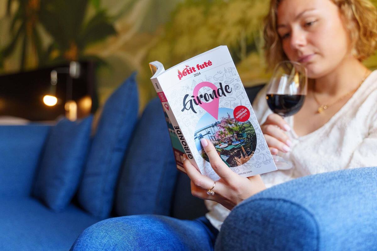 A person holding a book titled 'Gironde' and a glass of red wine, seated on a blue couch with a blurred background.