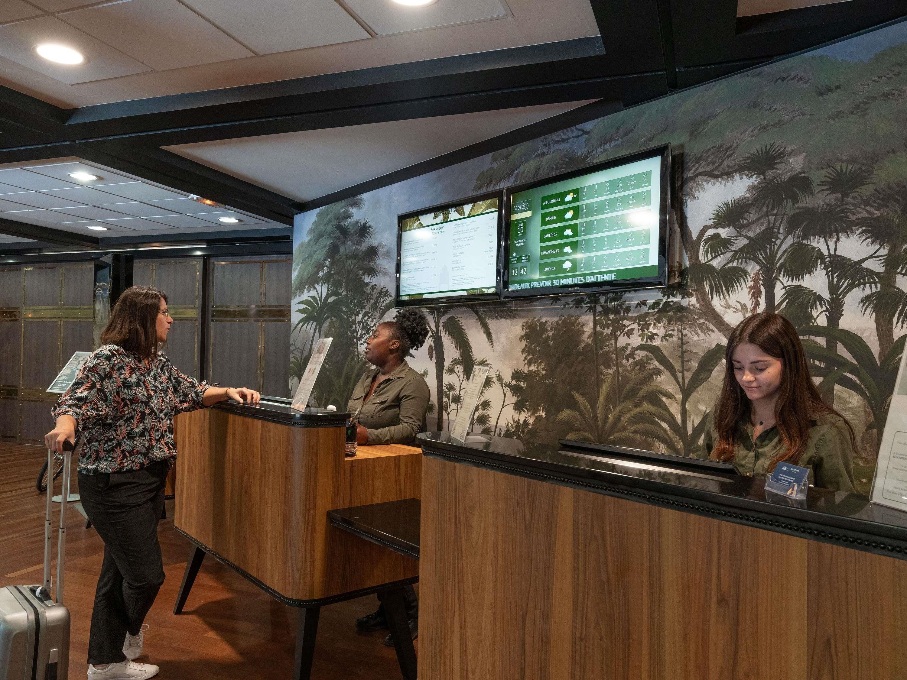 Un comptoir d’accueil doté d’un écran numérique, d’un plan de travail en bois et d’une fresque représentant des arbres tropicaux.